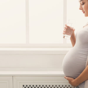 Pregnant woman drinking water at window Pregnant woman drinking water, standing at window. Young expectant lady have rest at home with glass, copy space. Pregnancy, healthcare, thirst concept